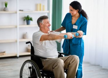 Female physiotherapist helping elderly man in wheelchair do exercises with dumbbells at health centre. Senior male petient undergoing rehabilitation after illness or injury at fitness clinic